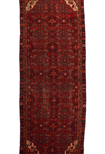 A traditional long runner rug with a deep red field, intricate geometric and floral motifs in black, beige, and rust, and a dark border.